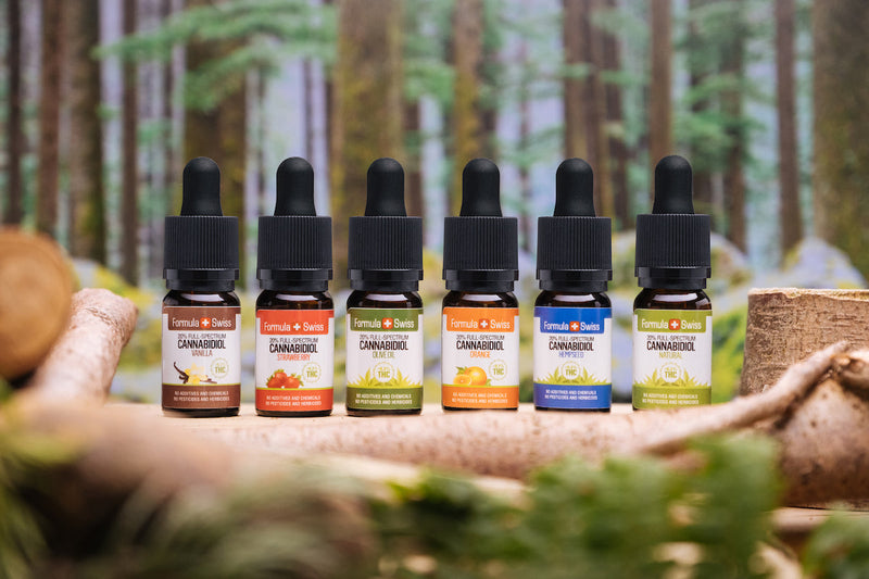 Șase uleiuri CBD Formula Swiss cu spectru complet în diferite uleiuri purtătoare și arome, în fața unei păduri